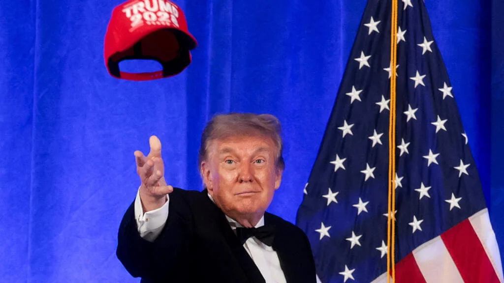 Дональд Трамп в смокинге подбрасывает красную кепку TRUMP 2028 у флага США