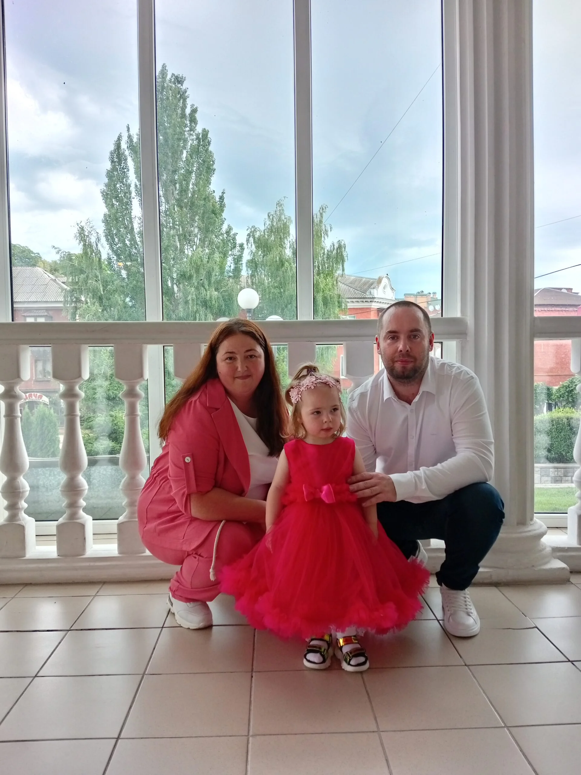 Родители с маленькой дочкой в красном платье позируют у больших окон
