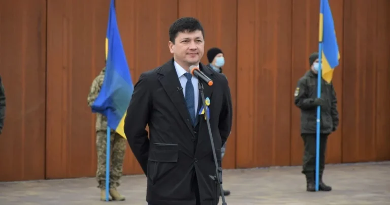 Виталий Ким выступает на фоне украинских флагов с военными