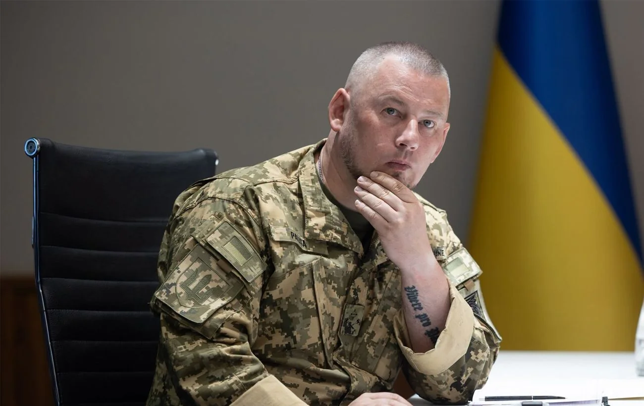 Павло Паліса в камуфляжі сидить за столом на тлі українського прапора
